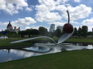 Giant spoon bridged over a reflective pond with a bright cherry perched on its tip.