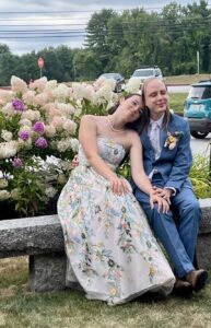 Photo of my son in a blue suit with his bride, who wears her wedding dress covered with stitched flowers. Her head is on his shoulder as they sit on a granite bench in front of hydrangeas.