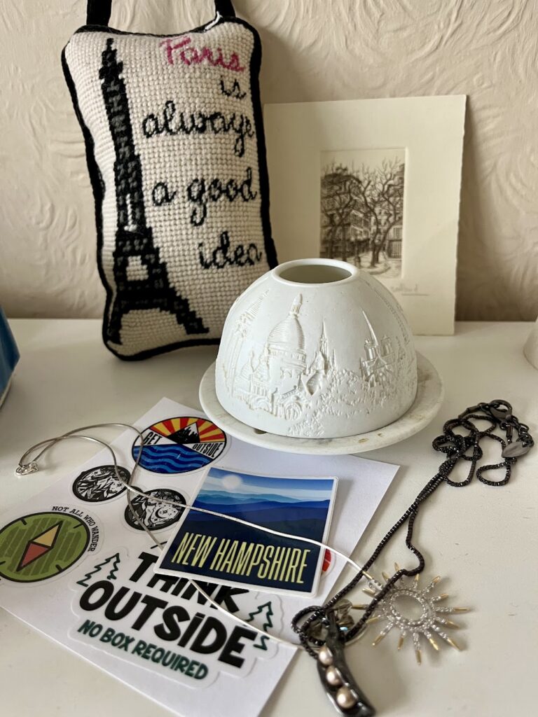 Collection of artefacts from our summer trip: my late aunt's sachet with the Eiffel Tower stitched on which reads 'Paris is always a good idea;' a small framed sketch of Rouen; a porcelain candleholder embossed with French landmarks; stickers portraying New Hampshire Mountains and with 'Go outside' slogans; a necklace with a glittering sunshine and one portraying pearlescent peas in a pod.