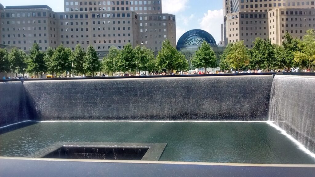 Memorial fountain in the footprint of the World Trade Centre, surrounded by people and trees and further on, more city buildings.